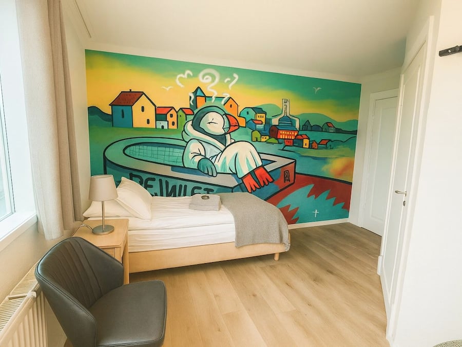A bedroom with a bed, a chair, a nightstand, a lamp, and a wall mural.