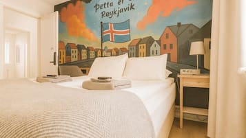 A hotel room with a bed, a nightstand with a lamp, and a mural on the wall depicting houses and a flag.
