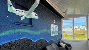 Ein Zimmer mit einem Wandgemälde eines Flugzeugs und einem Schild mit der Aufschrift 'FLATELY FLUGVOLLUR'. Vier Stühle und ein Fenster mit Blick auf Häuser sind vorhanden.