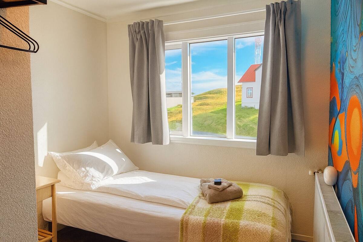 A bedroom with a bed, a window with curtains, a radiator, and a painting on the wall.