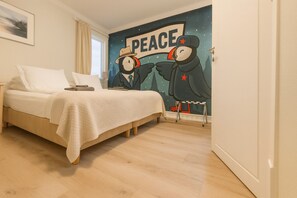 A bedroom with a bed, a nightstand, a wall mural depicting two penguins holding hands with a 'PEACE' sign, and a window with curtains. - Stykkisholmur Inn (Stykkisholmur)