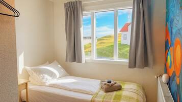 Una cama individual con colchón y almohadas blancas, una ventana con cortinas y un radiador en la pared.