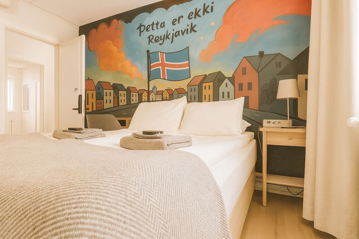 Quarto de hotel com cama, mesa de cabeceira com abajur e um mural na parede retratando casas e uma bandeira.