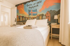 Una camera d'albergo con un letto, un comodino con una lampada e un affresco raffigurante case e una bandiera.