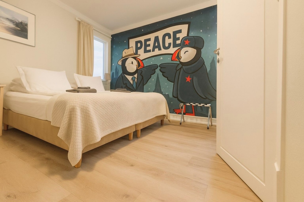 A bedroom with a bed, a nightstand, a wall mural depicting two penguins, and a window with curtains.