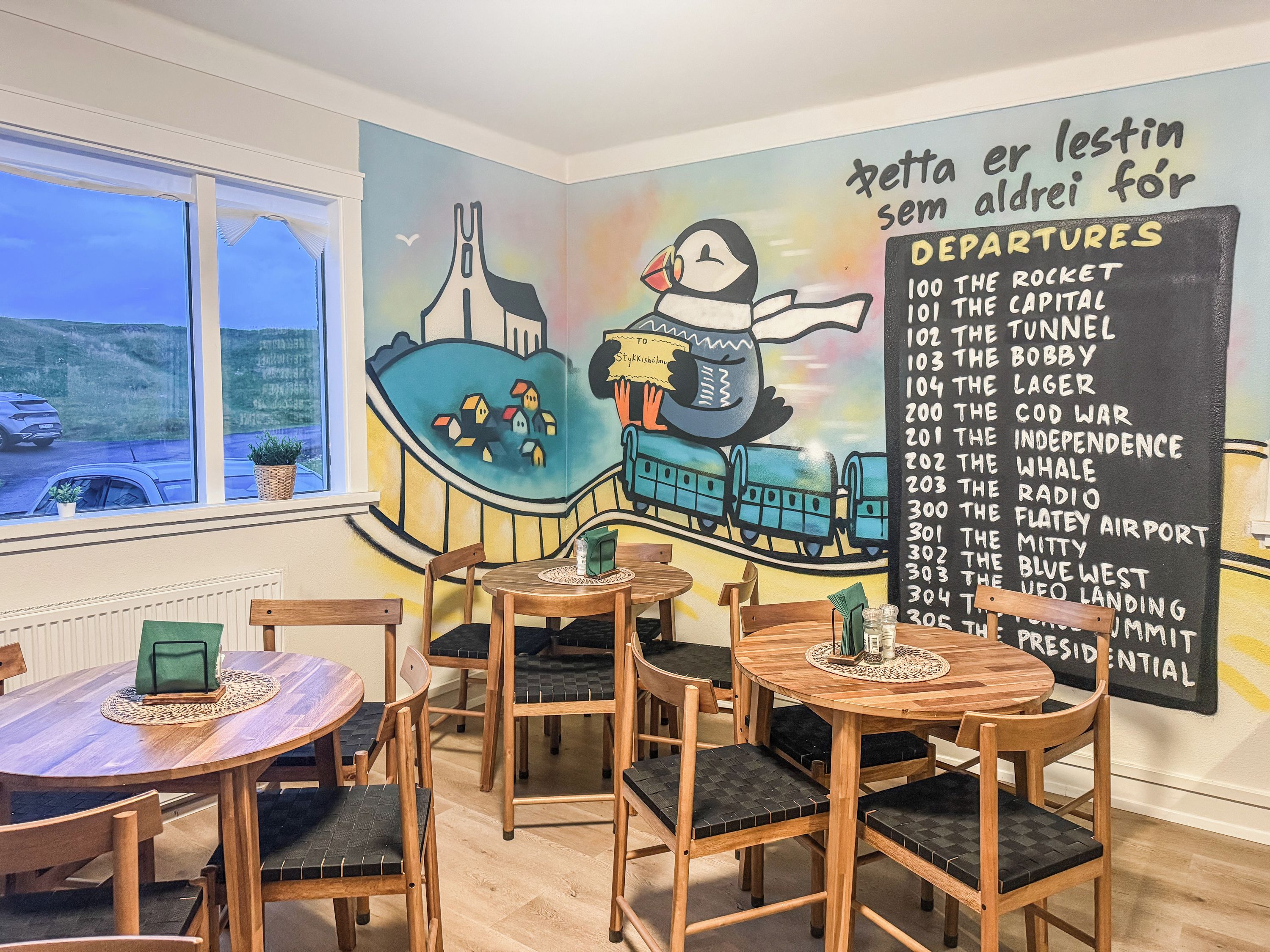 Ein gemütliches Restaurant mit Holztischen und -stühlen, einem Wandgemälde mit Zug und Häusern sowie einer Kreidetafel mit Abfahrtsorten.