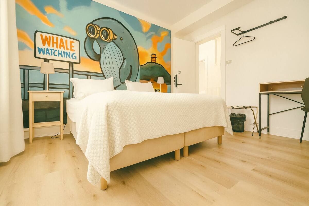A bedroom with a bed, a nightstand, a desk, and a chair. There is a mural on the wall depicting a whale watching scene.