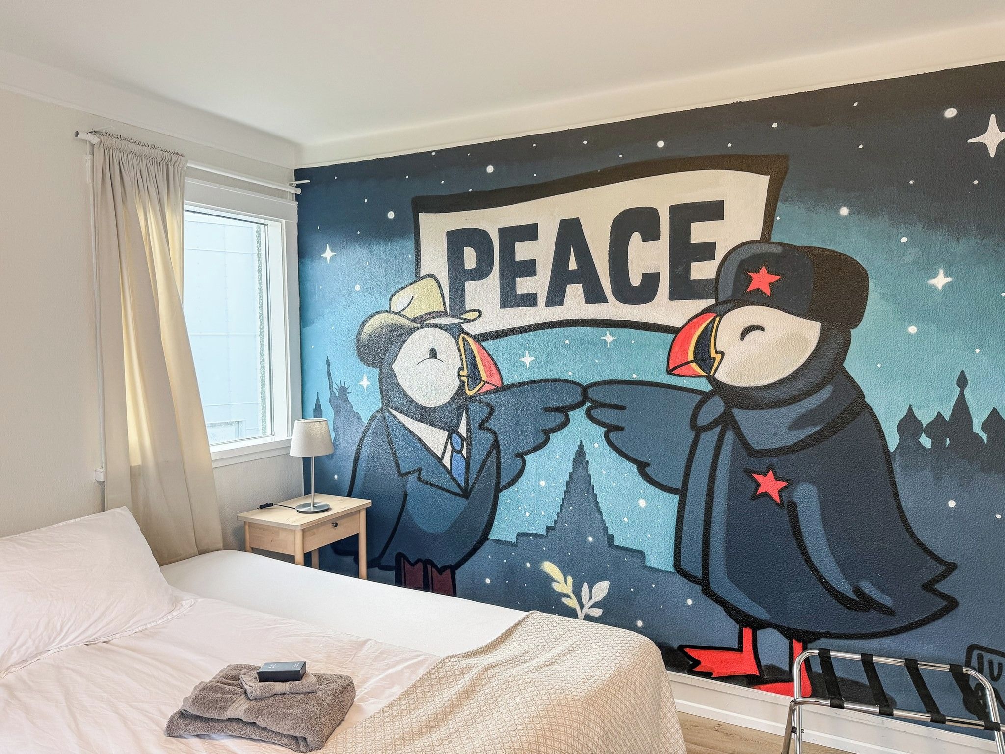 Une chambre d’hôtel avec un lit, une table de chevet, une lampe et une fresque murale représentant deux oiseaux et le mot « PAIX ».