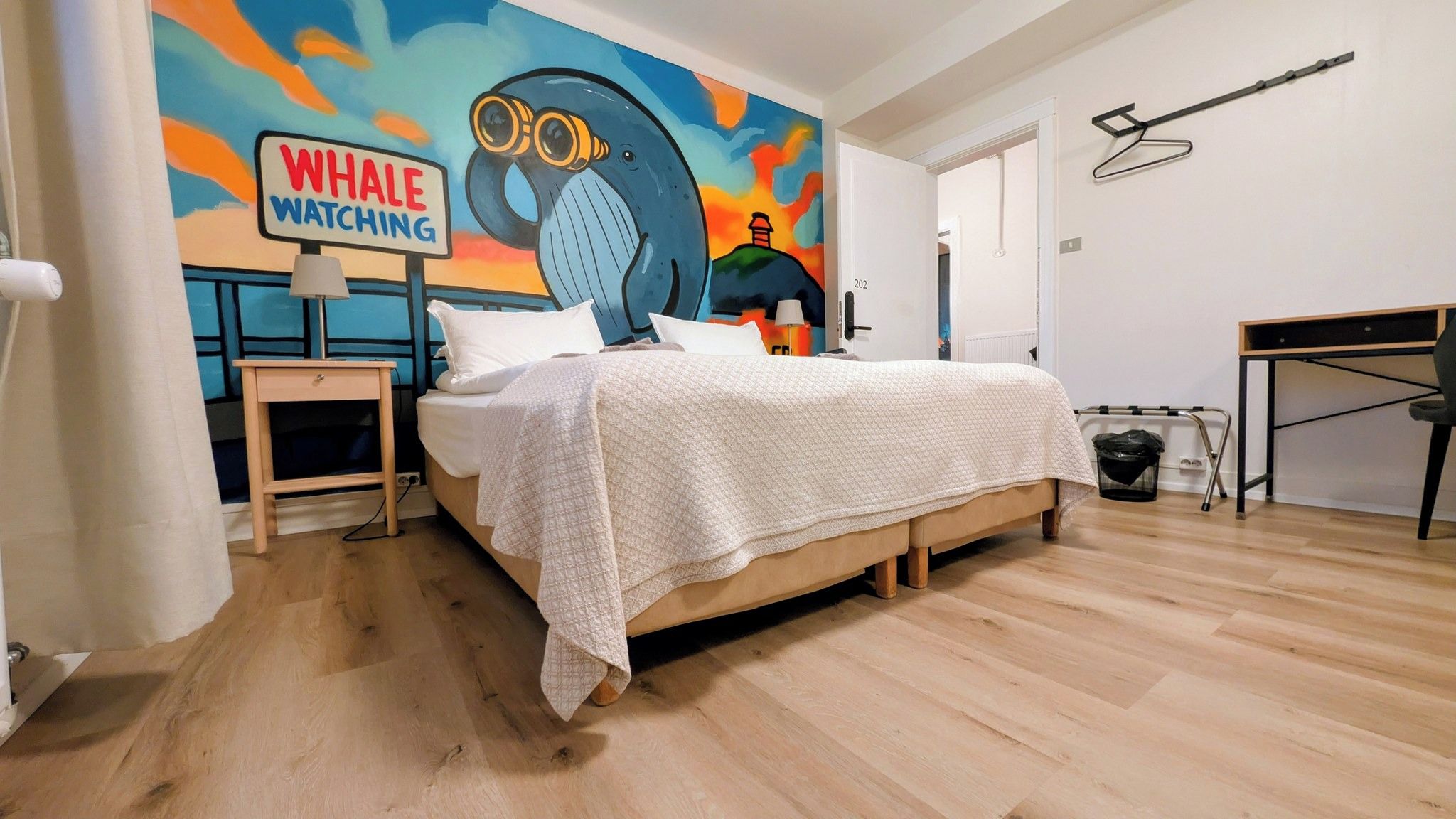 Quarto com cama, mesa de cabeceira, escrivaninha e parede com um mural colorido.