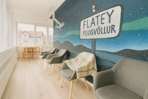 Un'area d'attesa con un murale, sedie grigie, un tavolo e un cartello con la scritta "FLATEY FLUGVÖLLUR".