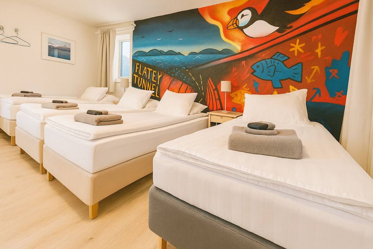 A hotel room with three single beds, each with a pillow and a rolled towel. The room has a large window with a view of the sea and a colorful mural on the wall.