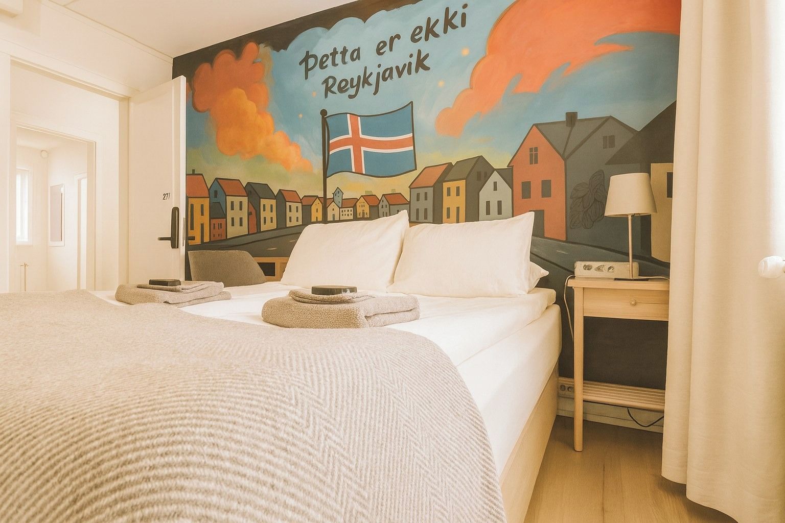 A hotel room with a bed, a nightstand, a lamp, and a wall mural depicting a town scene with a flag.