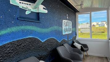 Una habitación con un mural de un avión y un cartel que dice "FLATELY FLUGVOLLUR". Hay cuatro sillas y una ventana con vista a casas.