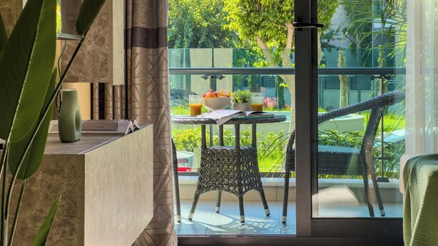 A modern living room with a large glass door leading to a balcony. The room features a marble side table with a vase and a book, and a wicker chair on the balcony. - Qoople Legends (Alanya)