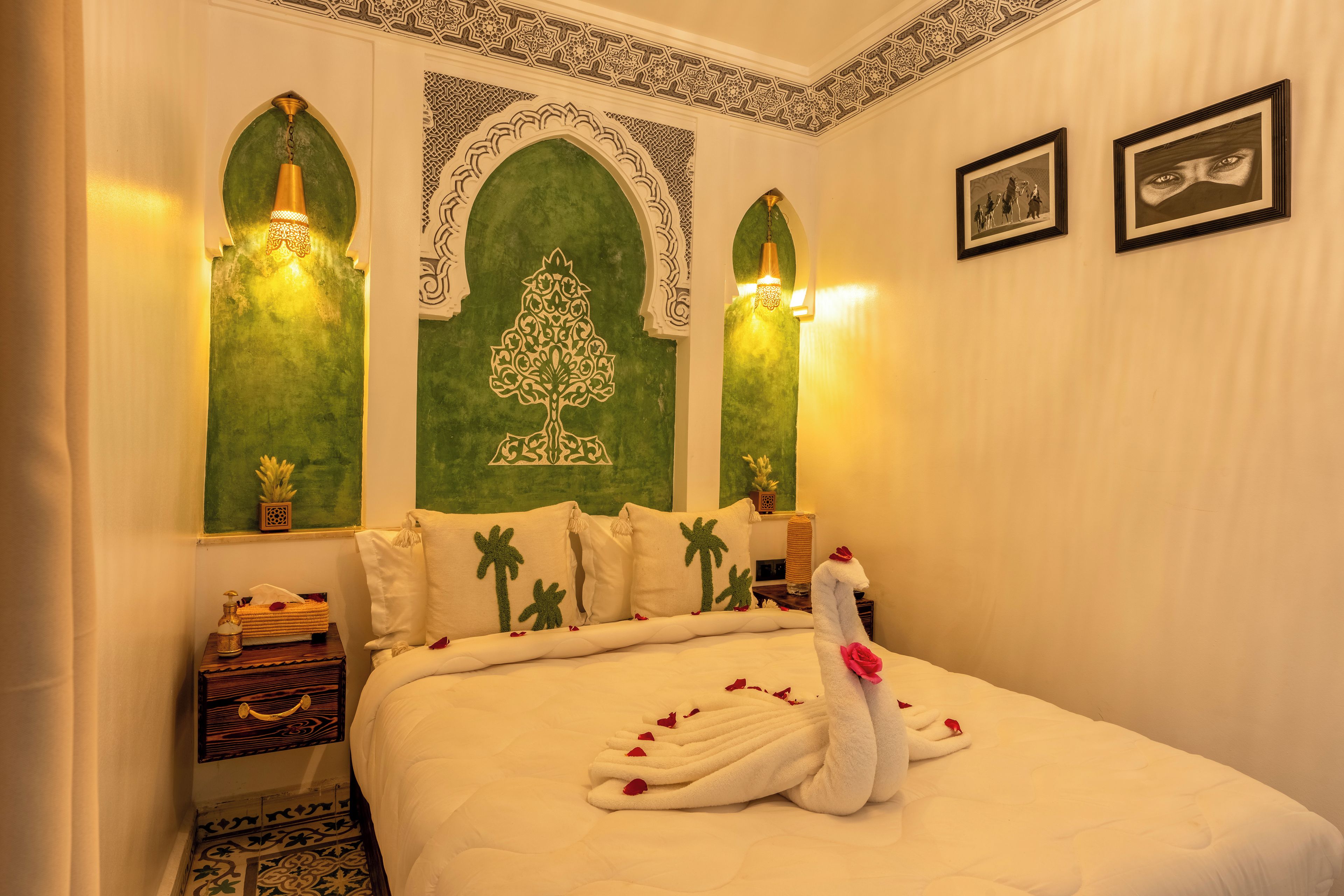 A neatly made bed with a swan decoration, a green wall with a tree design, and two wall-mounted lights.
