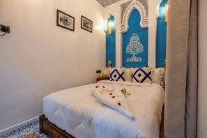 A neatly made bed with a white duvet and decorative pillows, a wall-mounted light fixture, and a framed picture on the wall. - Riad La Perle Bleue (Marrakech)