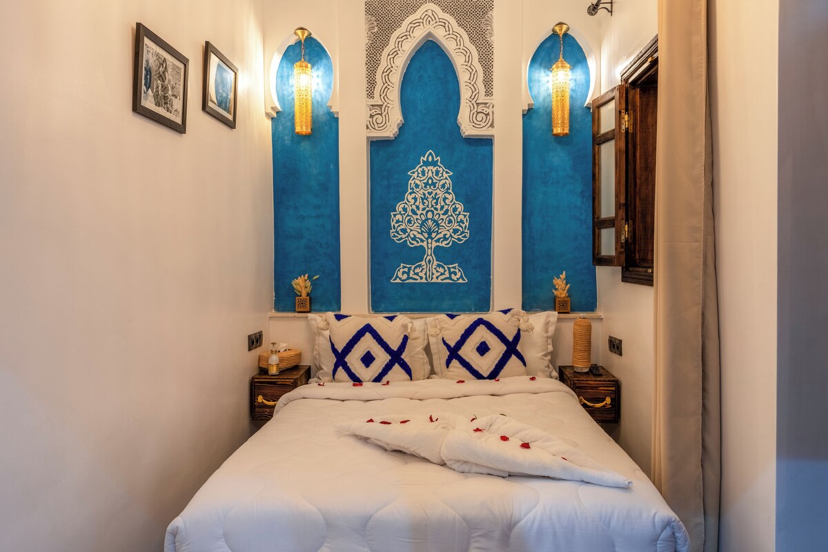 A neatly made bed with white linens and decorative pillows, a wall with a blue decorative panel featuring a tree design, and two wall-mounted lamps.