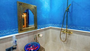 Casa de banho com parede azul, espelho com moldura dourada, tigela de vidro com pétalas de rosa, chuveiro manual e suporte para rolo de papel higiénico.