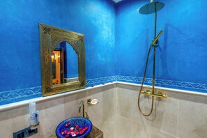 A bathroom with a blue wall, a gold-framed mirror, a glass bowl with rose petals, a handheld showerhead, and a toilet paper holder. - Riad La Perle Bleue (Marrakech)