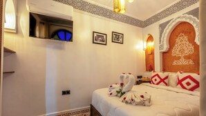 Quarto de hotel com cama, televisão de tela plana, padrões decorativos na parede e um candelabro.