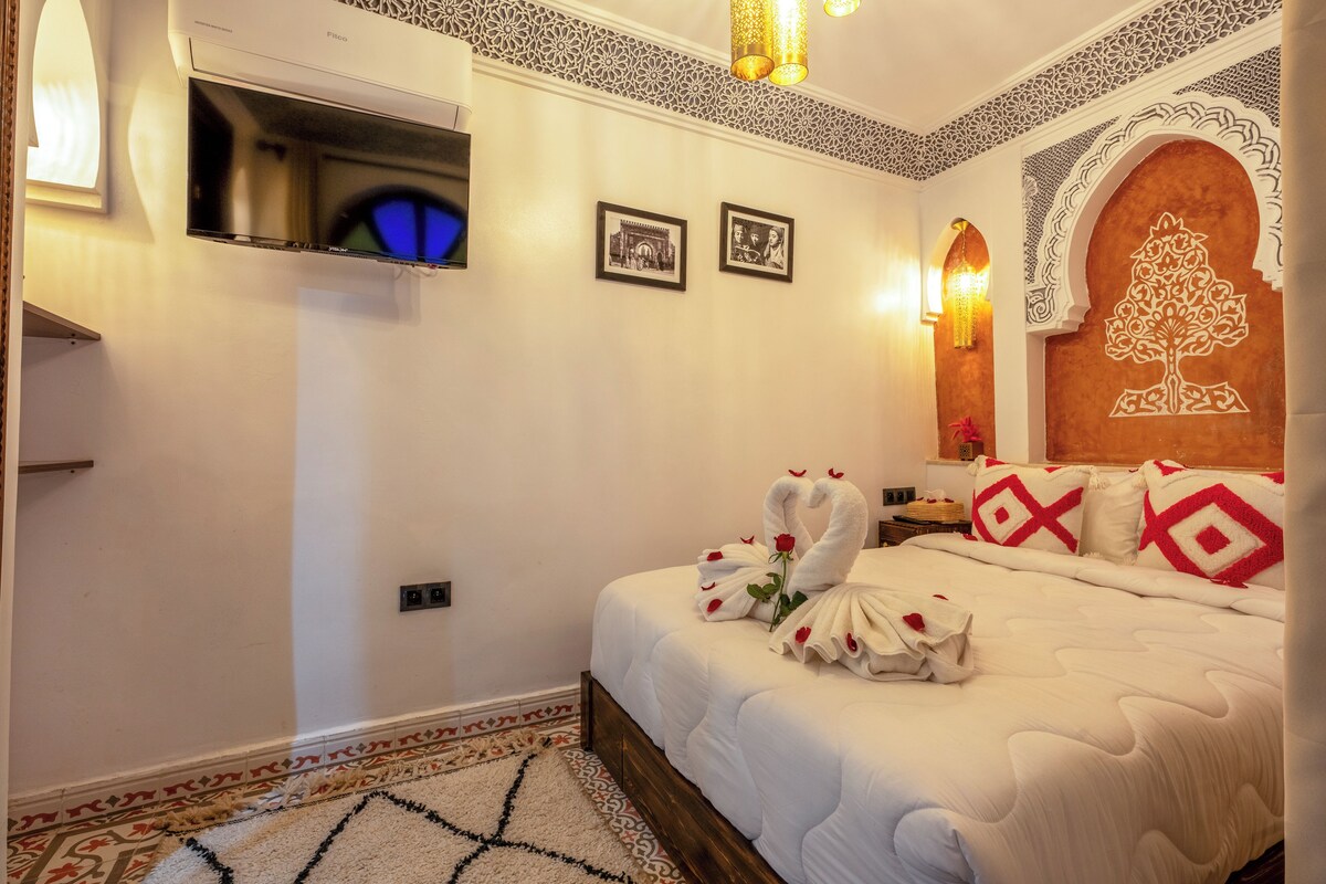 Quarto de hotel com cama, televisão de tela plana, padrões decorativos na parede e um candelabro.