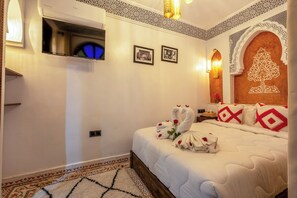 Quarto de hotel com cama, televisão de tela plana, padrões decorativos na parede e um candelabro.