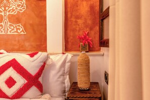 A cozy room with a patterned throw blanket, a decorative vase with pink flowers, and a textured wall art piece. - Riad La Perle Bleue (Marrakech)