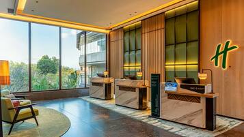 A modern hotel lobby with a reception desk, seating area, and large windows offering a view of greenery.