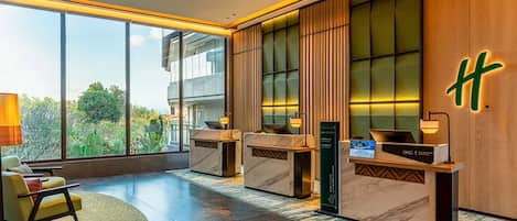 A modern hotel lobby with a reception desk, seating area, and large windows offering a view of greenery.