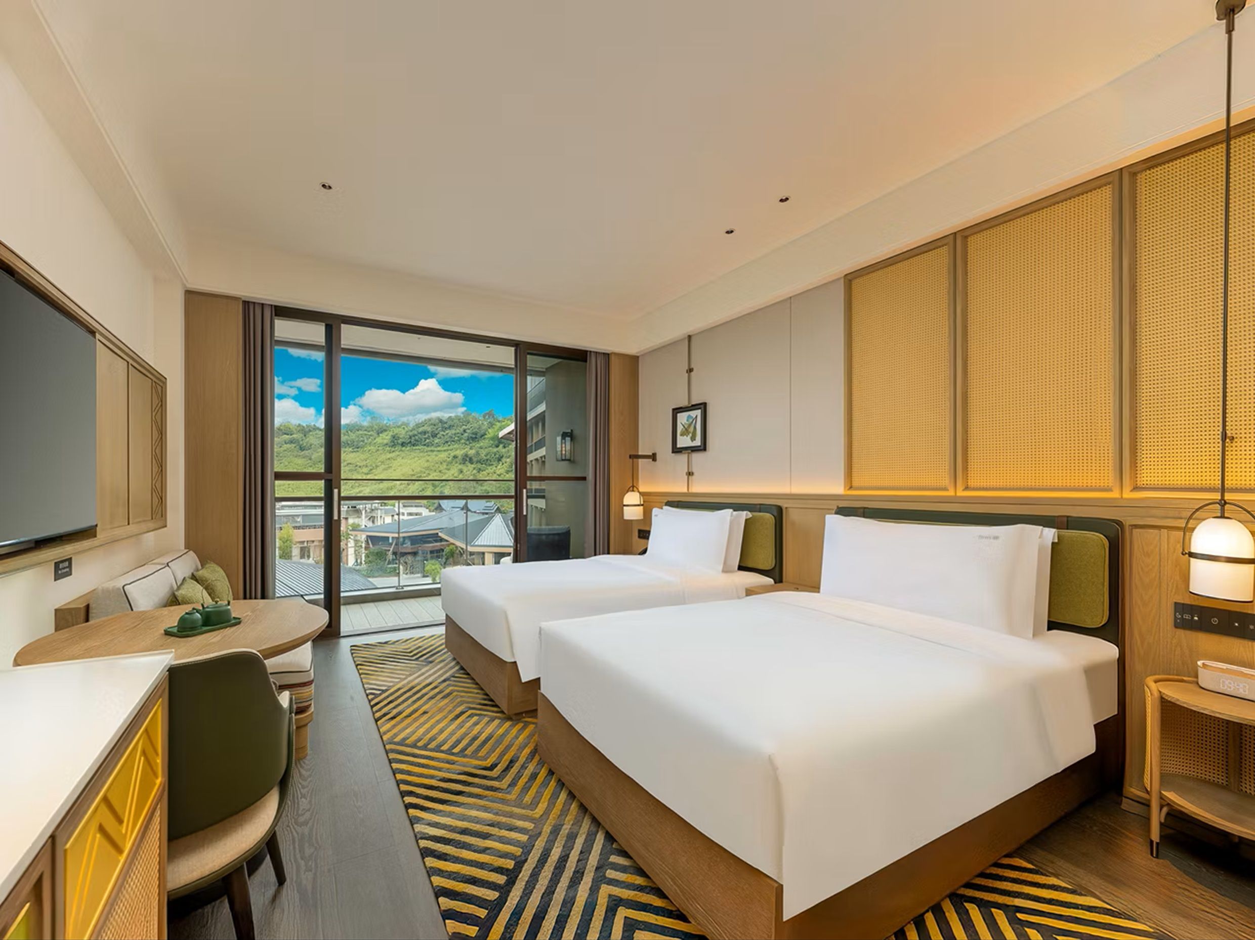 A hotel room with two beds, a desk, a chair, and a view of a green landscape.