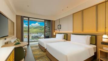 A hotel room with two beds, a desk, a chair, and a view of a green landscape.