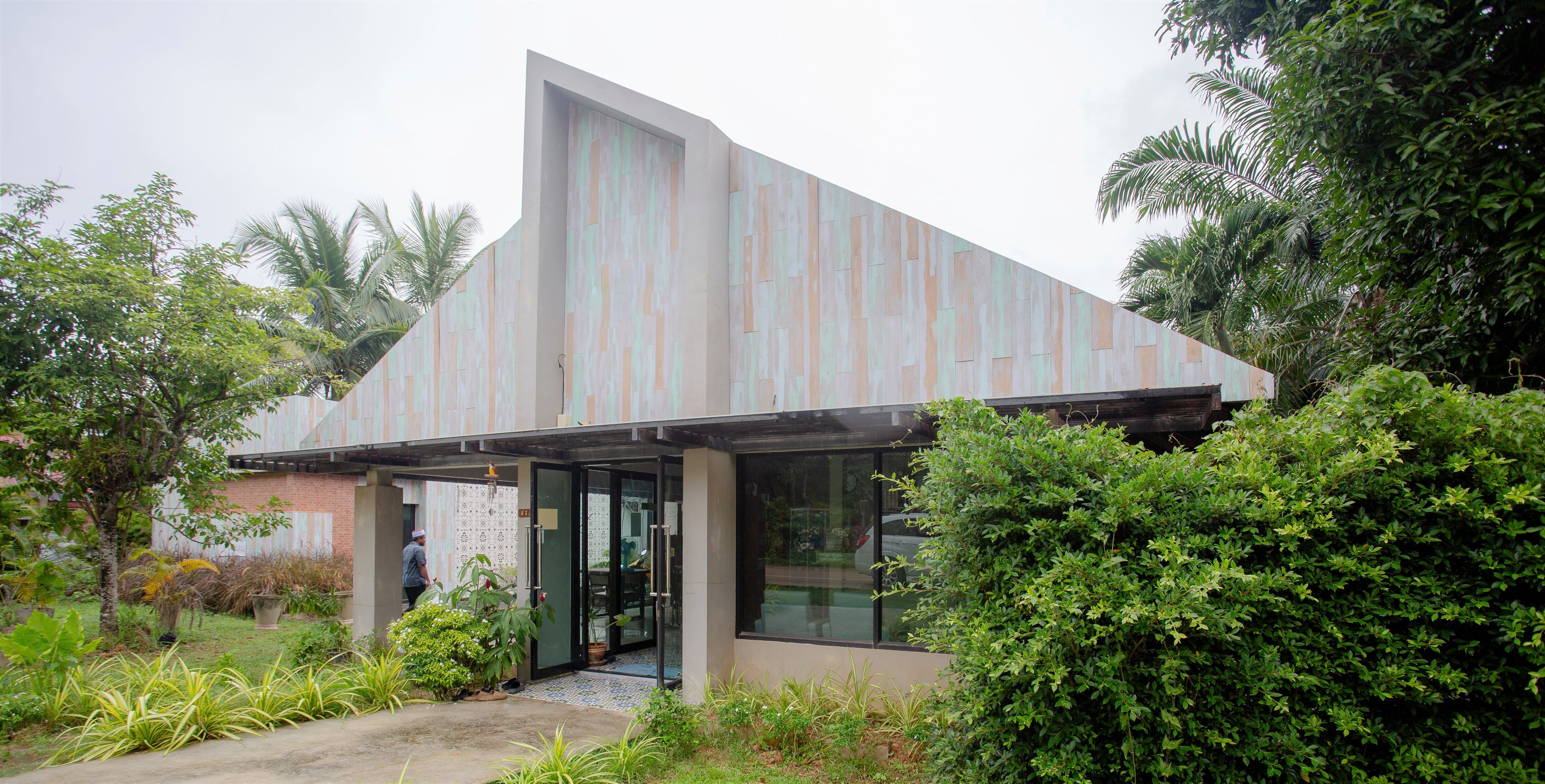A modern building with a sloped roof and large glass doors, surrounded by lush greenery.
