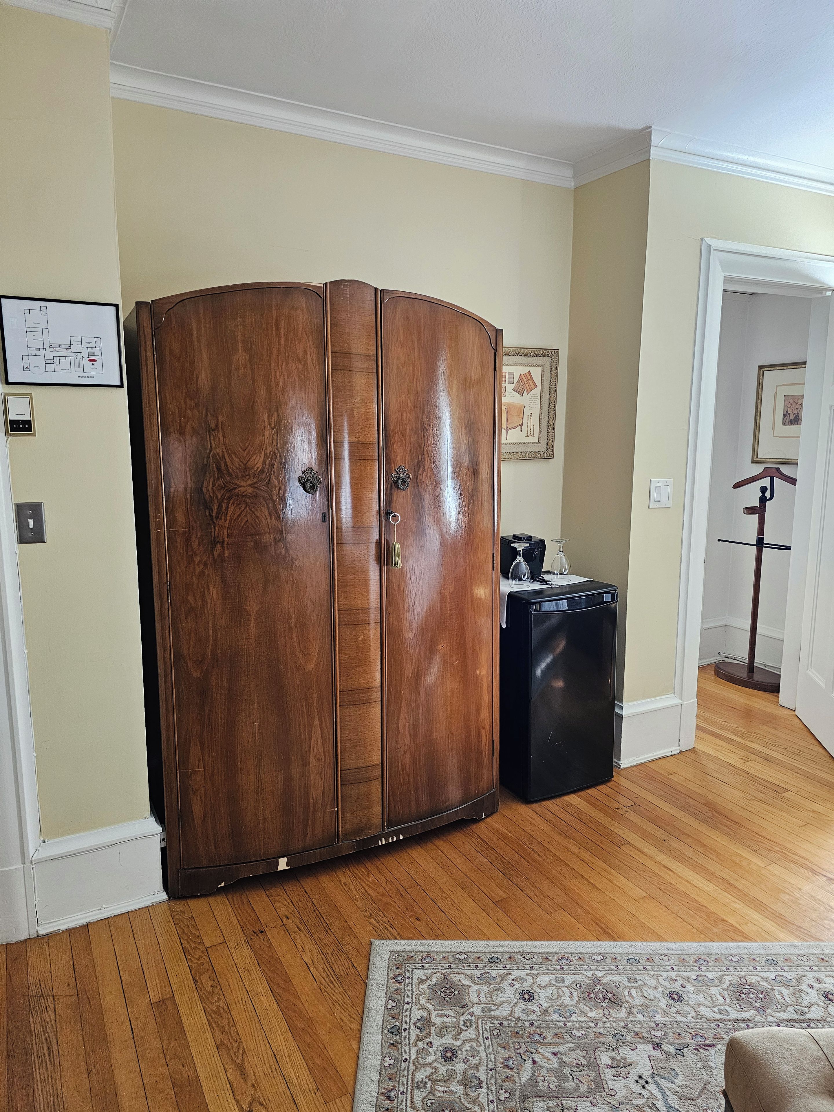 A room with a wooden wardrobe, a black suitcase, a patterned rug, and a door leading to another room.