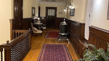 A hallway with wooden flooring, a red patterned rug, a staircase with a wooden railing, and a wall-mounted light fixture.