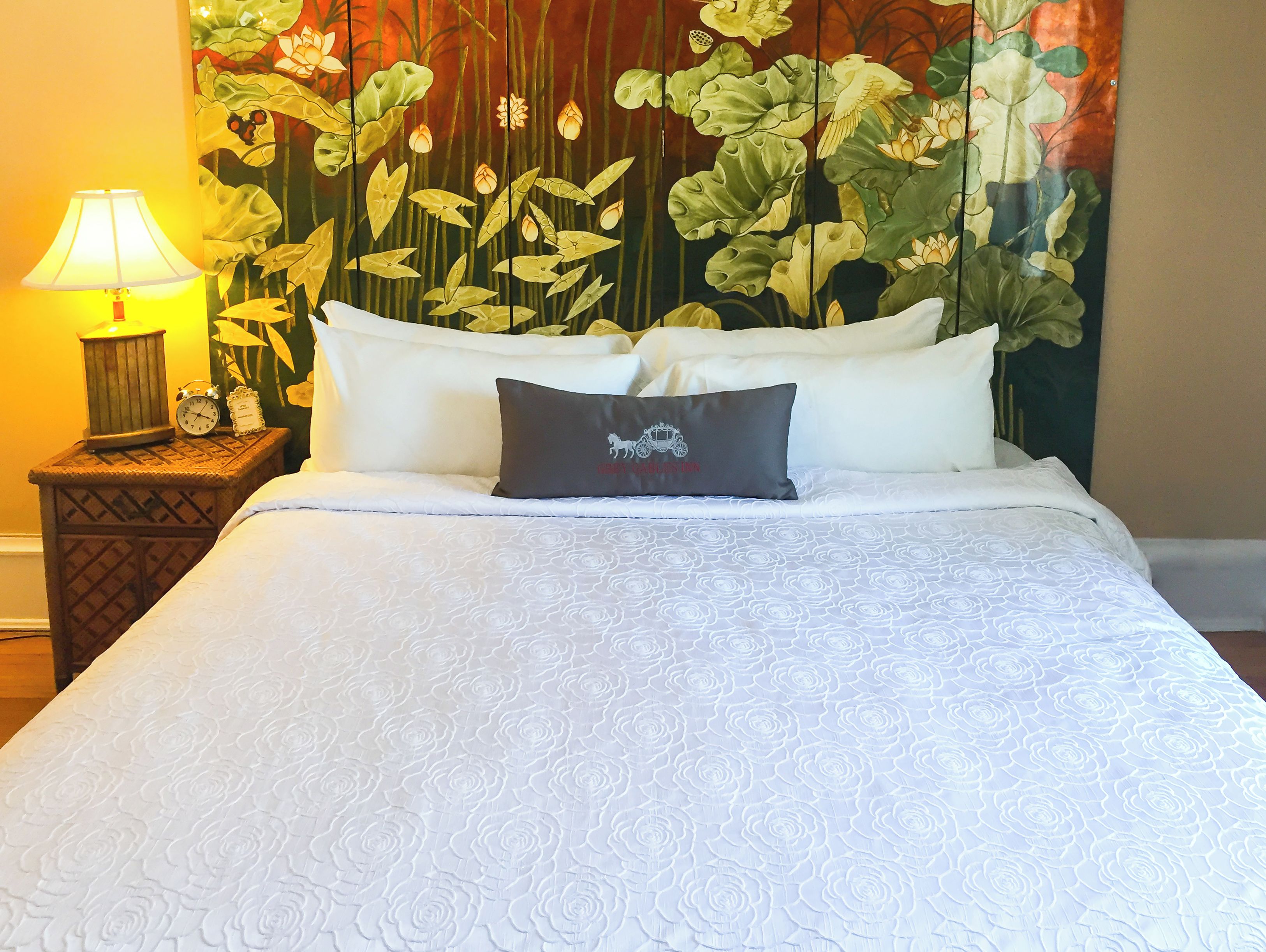 A neatly made bed with a white rose-patterned duvet, two pillows, and a decorative pillow with a horse logo. A lamp and a clock are on a wooden nightstand beside the bed.
