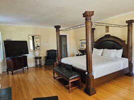 Una cama con dosel y tablero de madera, un televisor de pantalla plana, un sillón de cuero y un banco en una habitación de hotel con piso de madera.