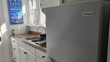 A kitchen with a white refrigerator, a microwave, and a sink. There is a window with curtains and a blue sign on the wall.