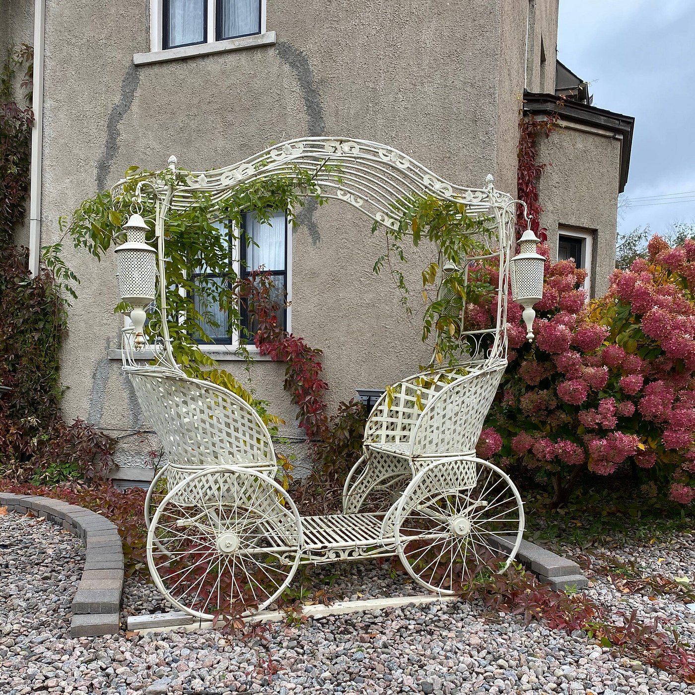 A white metal carriage with a basket, positioned in front of a house with a window and a door.