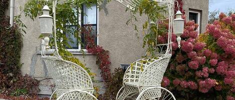 Une petite voiture en métal blanc, dotée d’un panier, est placée devant une maison qui a une fenêtre et une porte.