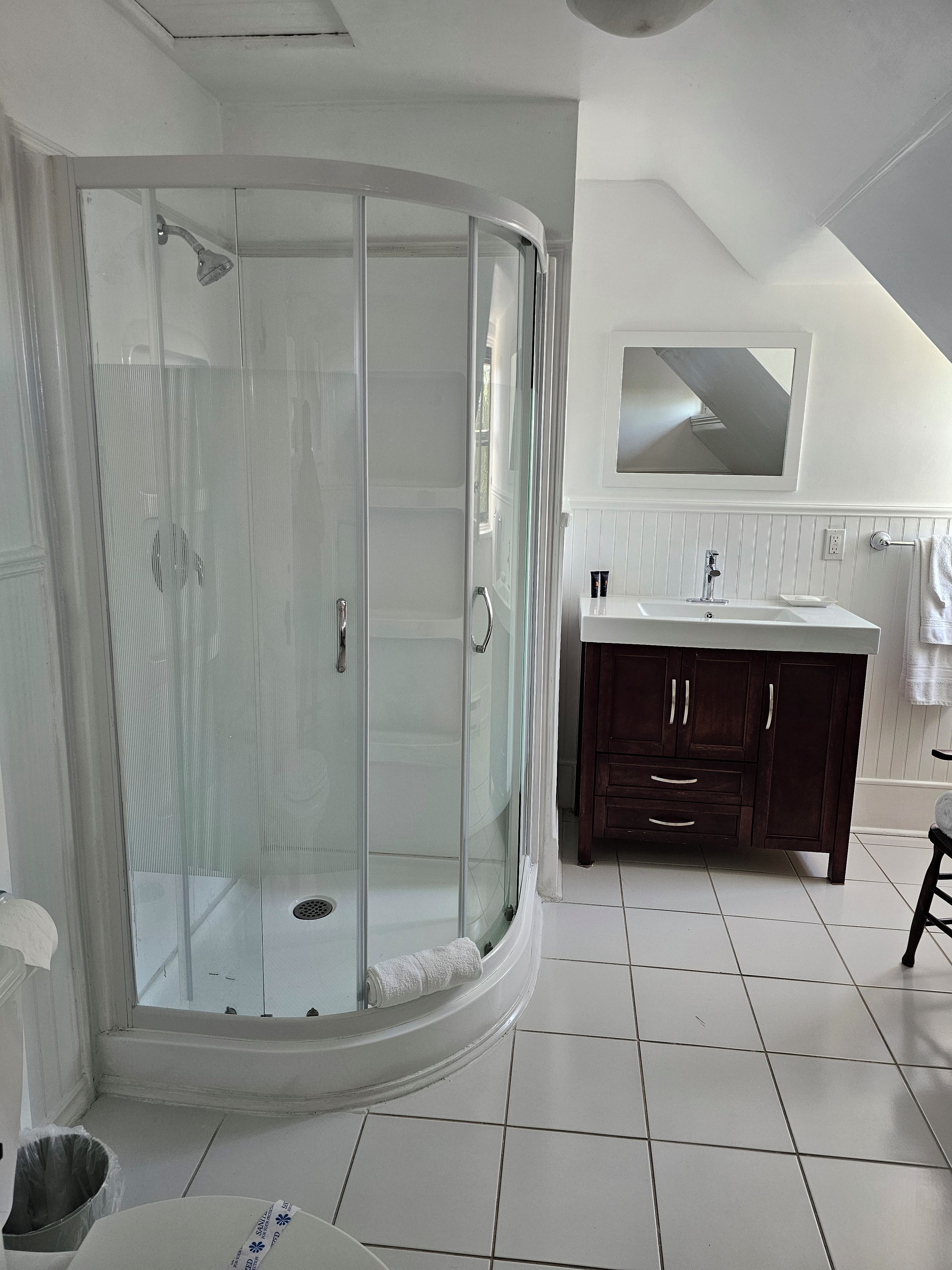 Un baño moderno con ducha envuelta en vidrio, azulejos blancos, tocador de madera oscura y un espejo grande.