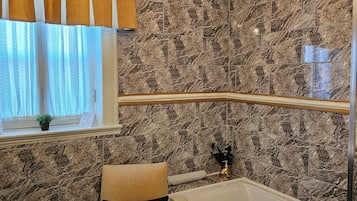 Une salle de bain avec un mur en marbre, une baignoire, une chaise, un lustre et une fenêtre avec des rideaux.