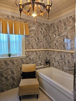 Un baño con pared de mármol, una bañera, una silla, un candelabro y una ventana con cortinas.