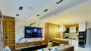 A modern living room with a flat-screen TV, a sofa, a coffee table, and a kitchen area in the background. - The GlobalNomad Enterprise (Lekki)