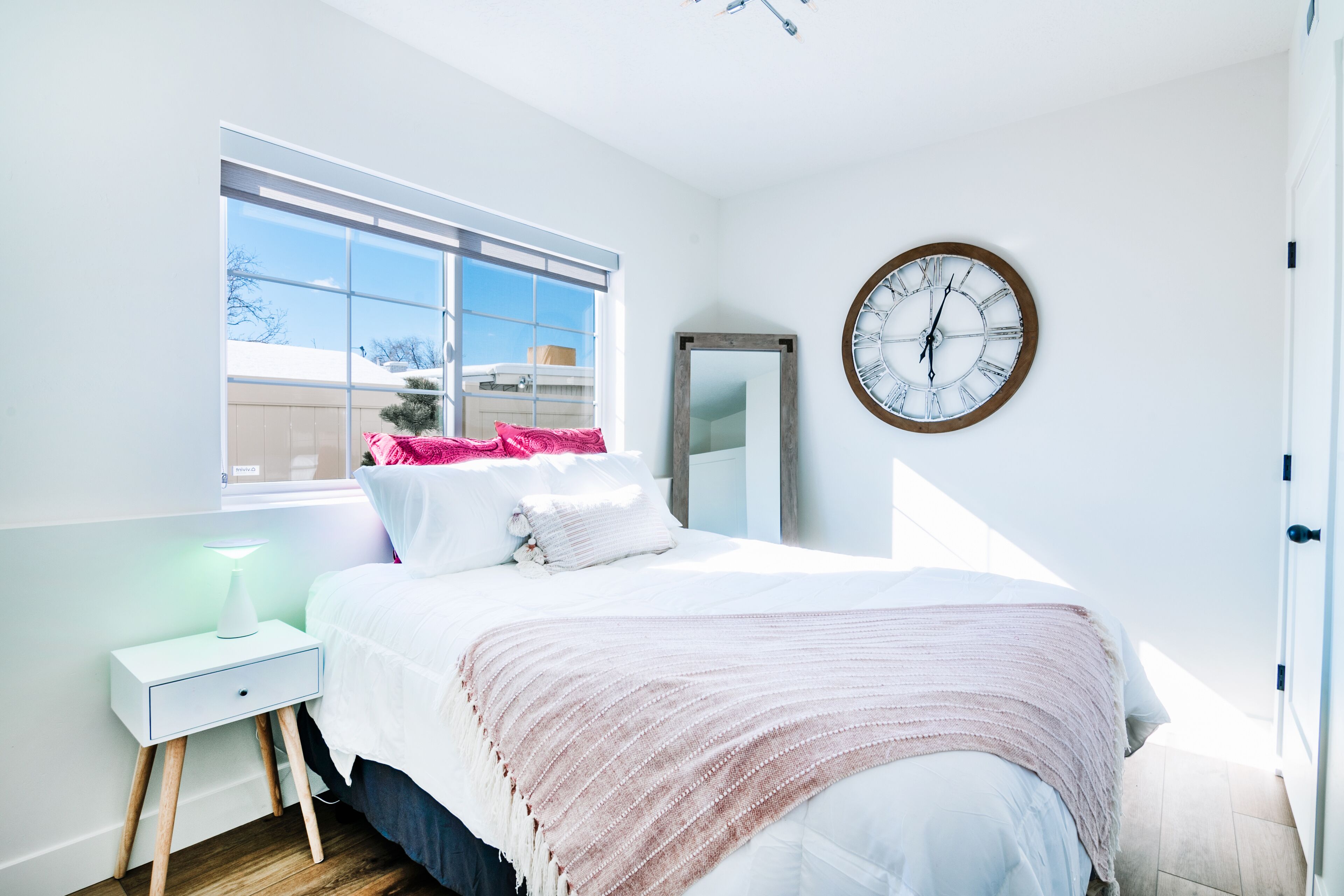 A bedroom with a bed, a bedside table with a lamp, a large window, and a wall clock.