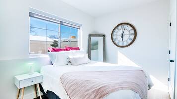 A bedroom with a bed, a bedside table with a lamp, a large window, and a wall clock.