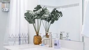 Ein Badezimmer mit weißem Waschbecken, einem Spiegel, einer Duschkabine und zwei Topfpflanzen.