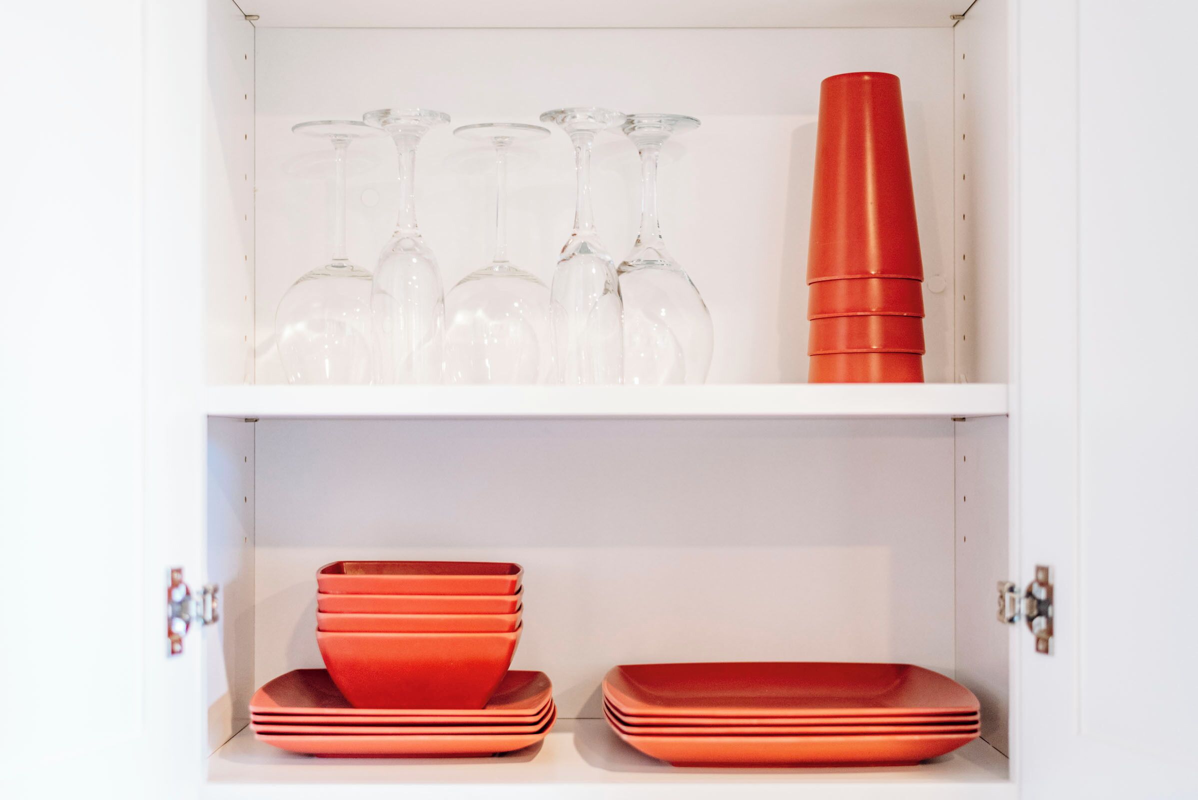 A white cabinet with two shelves. The top shelf holds several clear wine glasses and a tall red object. The bottom shelf contains several red plates and bowls.