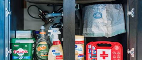 Un armario de almacenamiento con productos de limpieza, incluyendo detergente Cascade, jabón para platos Gain, toallitas desinfectantes y un botiquín de primeros auxilios.