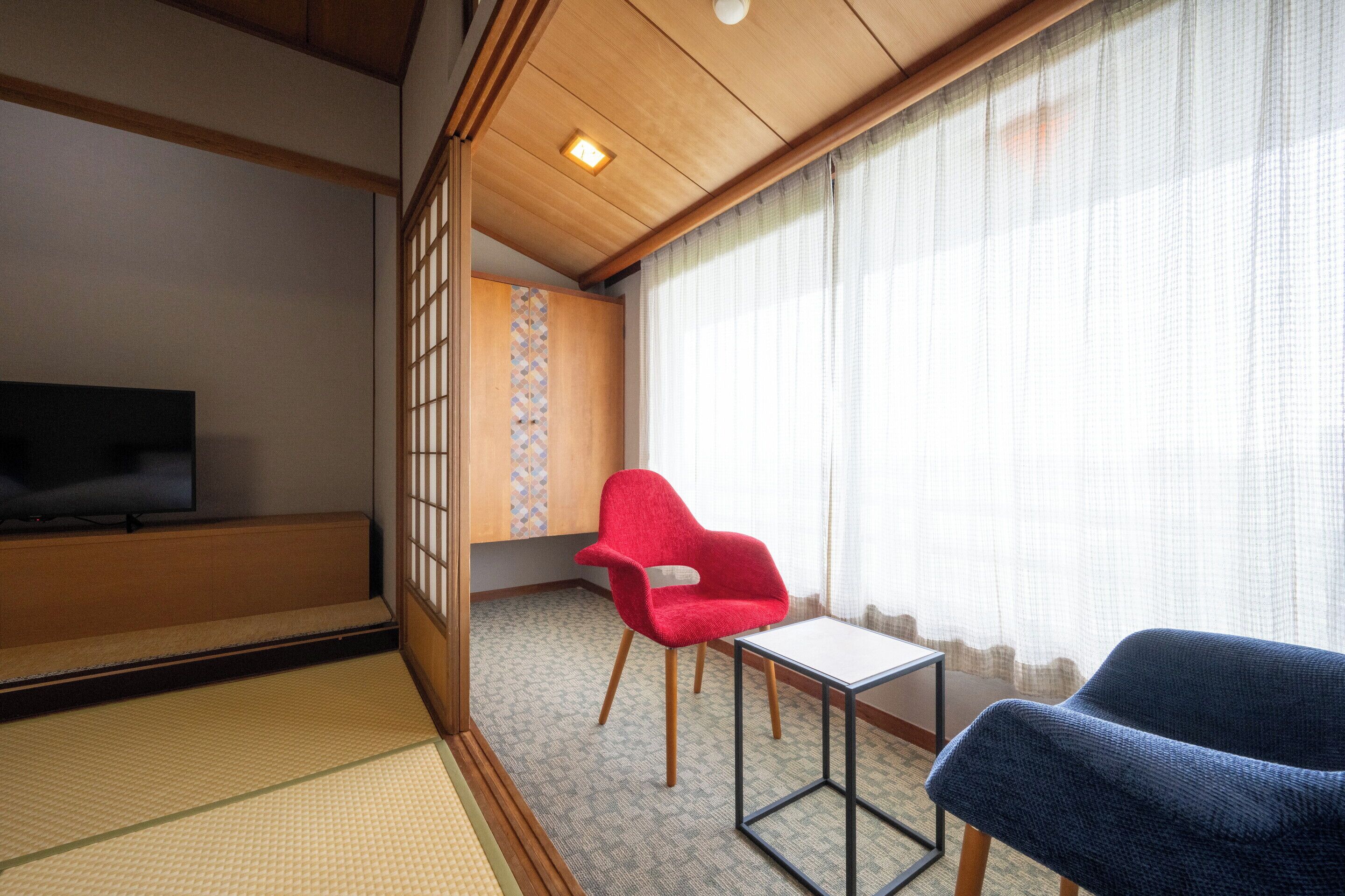 A traditional Japanese room with tatami flooring, a television, a red chair, a blue chair, and a small table.