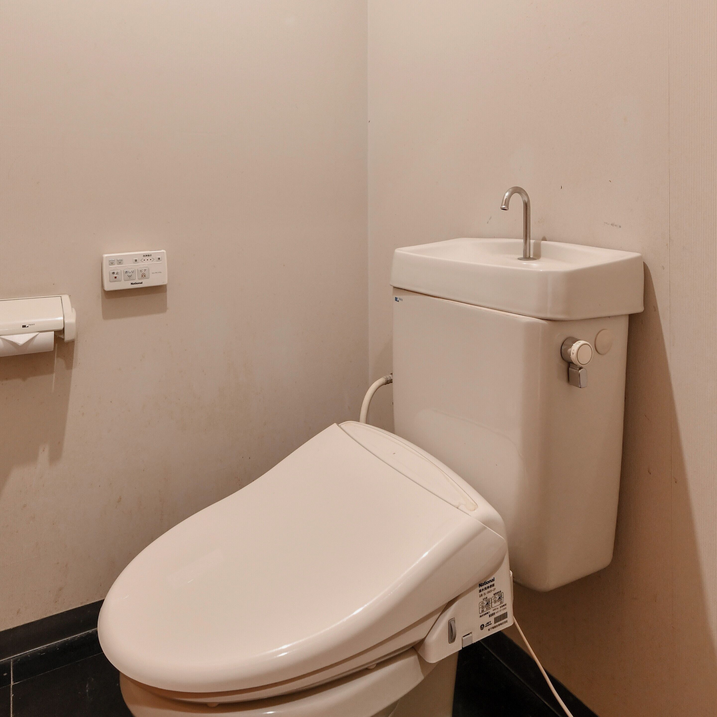 Un inodoro con asiento y tanque blancos, un pulsador de descarga en la pared y una tubería visible.